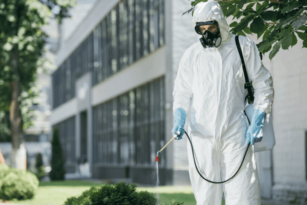 An urban pest management technician wearing personal protective equipment sprays pesticide outside an office building.